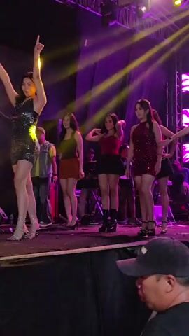 Four young women are dancing on a stage under bright lights. They are all wearing short, sequined dresses and high heels. The woman on the far left is wearing a black dress, the woman next to her is wearing a burgundy dress, the woman in the middle is wearing a red top and a black skirt, and the woman on the far right is wearing a dark purple dress. There are several people in the background, including musicians and crew members. The stage is set up with equipment, including speakers and keyboards. The overall atmosphere appears to be a lively performance.