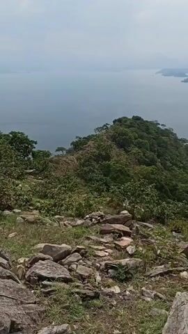 The video shows a view from a rocky, grassy hillside overlooking a large body of water. The hillside is covered with green vegetation. In the distance, several small islands can be seen in the water. The sky is overcast. The camera pans slightly to the right, providing a wider view of the landscape.