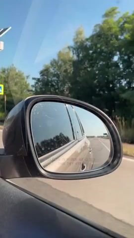 A view from inside a vehicle shows a side mirror reflecting the road and scenery behind the car. The mirror is slightly cracked. A blue sign with a yellow pedestrian symbol is visible in the distance, along with trees and a clear blue sky. The vehicle appears to be moving on a paved road.