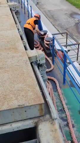 A person wearing a hard hat and an orange vest is securing a thick rope to a bollard on the deck of a vessel. The rope is pink and coiled on the deck. The person is actively pulling and wrapping the rope around the bollard. In the background, there is a blue railing, a concrete embankment, and green grass. The scene appears to be a port or a docking area.