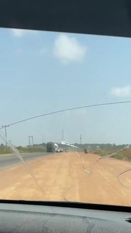 From the perspective of inside a vehicle with a cracked windshield, the video shows a convoy of various vehicles moving on a two-lane road under a clear, pale blue sky. There are several large trucks, one with a canvas covering and another that appears to be carrying a large tank. Smaller vehicles, including vans and cars, are interspersed within the convoy. The road has an unpaved shoulder and is lined with low vegetation. Transmission towers are visible in the distance. The vehicles are moving along the road, kicking up dust.