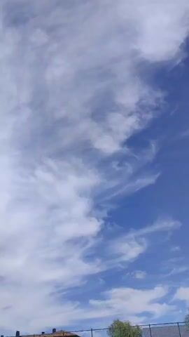 The video shows a time-lapse of clouds moving across a blue sky. In the foreground, there are trees and a fence. There are also utility poles visible. The clouds are white and wispy, and they appear to be moving from left to right. The sky is a bright blue, and the sun is shining. The video is shot from a low angle, looking up at the sky.