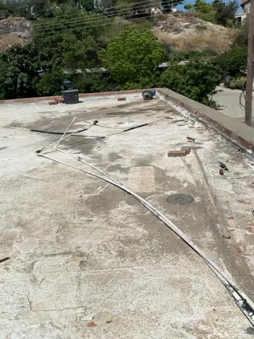 The video shows a rooftop with a concrete surface and several pipes running across it. There are also some bricks and a red bucket filled with gray material, likely cement, near the edge of the roof. A new brick wall is being constructed along the edge of the roof, with fresh cement visible. In the background, there are houses, trees, and a road. The overall scene suggests ongoing construction or repair work on the rooftop.