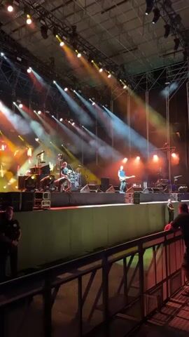 A live band performs on a brightly lit stage during a concert. The stage is equipped with drums, amplifiers, and various musical instruments. The band members are visible on stage, with one person playing the guitar and another at the drums. Smoke or fog machines are being used to create atmospheric effects, with beams of colored light illuminating the stage from above. In the foreground, a crowd of people watches the performance from behind a railing. Security personnel are also visible, standing near the stage. The overall environment suggests an outdoor concert venue with a covered roof structure.