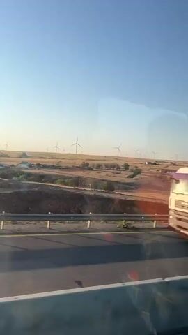 A white semi-trailer truck drives on a highway. The truck is large and has a white cargo container. It is moving from left to right in the frame. Reflected in the truck's window are trees and a clear blue sky. In the background, there is a landscape with rolling hills and several wind turbines. The sun is low in the sky, casting long shadows. The road has two lanes and is separated by a concrete barrier.