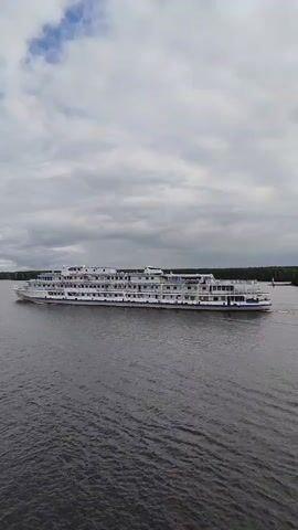 An aerial drone shot captures a large white passenger ship with multiple decks cruising on a dark body of water under a cloudy sky. The ship is angled slightly, showing its port side, and is moving from left to right. The water is calm, with a slight wake trailing behind the vessel. The background consists of a dark green, tree-lined shore on the right and a faint landmass on the left. The sky is filled with varying shades of gray clouds, with patches of white suggesting sunlight trying to break through. The drone maintains a steady altitude and position relative to the moving ship.