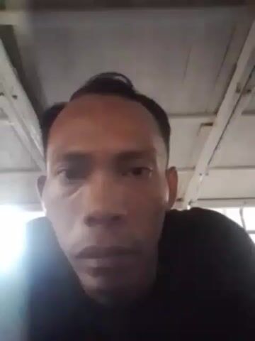 A close-up video of a man's face. He is wearing a black t-shirt. The man has dark hair and is looking directly at the camera. The background is out of focus and appears to be a wooden ceiling. The lighting is dim, with a bright light source coming from the left of the frame.