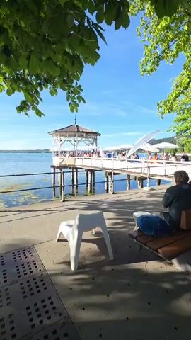 The video displays an outdoor scene beside a body of water on a sunny day. In the foreground, a white plastic chair sits on a concrete walkway. To the right, a person is seated on a wooden bench, facing away from the camera, with their legs crossed. Behind the bench, a large tree with green leaves partially obscures the view. Further back, a white wooden pier extends over the water, topped with a gazebo-like structure and several white umbrellas, suggesting an outdoor dining or seating area. People are visible on the pier, seated at tables. The water is calm with some aquatic vegetation visible near the shore. The sky is blue with scattered clouds. The overall atmosphere is serene and leisurely.