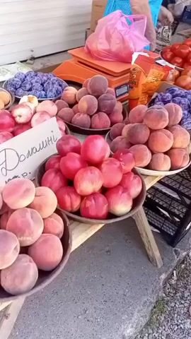 The video shows an outdoor market scene. In the foreground, a woman with long dark hair and sunglasses is standing at a fruit and vegetable stall, looking through her purse. She is wearing a green and white patterned dress. The stall displays a variety of produce, including peaches, plums, tomatoes, and red bell peppers. In the background, an older woman in a blue top is interacting with the stall owner. The market appears to be on a street with buildings and trees visible. The lighting suggests it is daytime.