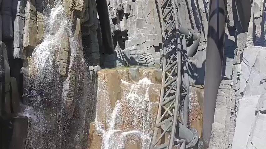 The video shows a roller coaster track weaving through a rocky, artificial landscape with waterfalls. Water cascades down rough-hewn rocks, creating a dynamic and visually appealing scene. The roller coaster tracks are a dark gray, contrasting with the lighter gray of the rocks and the white spray of the water. The overall impression is one of a thrilling amusement park attraction designed to immerse riders in a naturalistic, yet dramatic, environment.