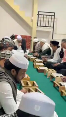Here is a concise description of the video:

The video shows a group of men sitting in rows on the floor in what appears to be a room with green carpeting. The men are dressed in religious attire, including various hats and robes, and most of them are reading from books on small wooden stands. The man in the front row on the left has a beard. A man in a suit is sitting at the head of the rows of men. There is a staircase behind them.