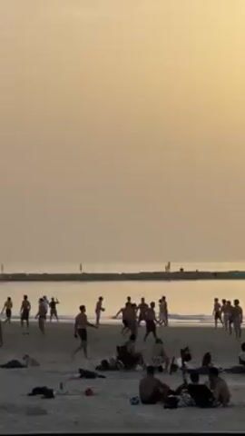 The scene is a beach at sunset. Many people are present. Some are playing with one or more soccer balls near the water’s edge, while others are sitting or lying down on the sand. The sky is a pale yellow color. There is a breakwater in the distance with a few people walking on it.