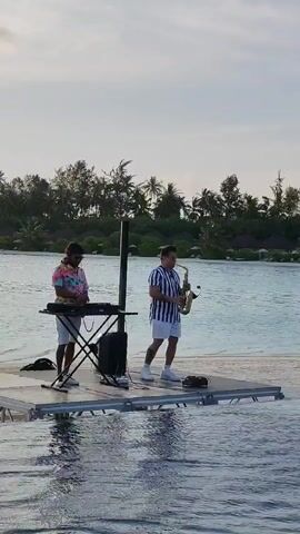 Two men are standing on a floating platform in the water. The man on the left is wearing a brightly colored floral shirt, white shorts, and headphones, and he appears to be operating a DJ setup with a keyboard. The man on the right is wearing a blue and white striped shirt, white shorts, and is playing a saxophone. A tall speaker stands between them. The background shows a shoreline with palm trees and some buildings, under a cloudy sky. The water is calm, and there are people swimming in the foreground. The platform is made of metal and wood.