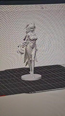 The video shows a 3D model of a female anime-style character rotating on a platform. The character has long, flowing hair, is wearing a revealing outfit, and is standing on a circular base. The platform appears to be a grid, suggesting it's a digital workspace for 3D modeling or printing. The background is a blurred grey texture.