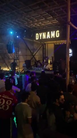The video shows a crowded indoor venue with a stage at the back where a DJ is performing. The venue has a rustic aesthetic with exposed wooden beams and a thatched roof. The lighting is dim and features various colored spotlights and lasers, creating a party atmosphere. A large sign above the DJ booth reads "DYNAMO" in illuminated letters. A diverse group of people, predominantly young adults, are seen dancing and socializing. Some individuals are in the foreground, facing the camera, while others are further back, interacting with each other or watching the DJ. The overall scene suggests a vibrant nightclub or bar setting.