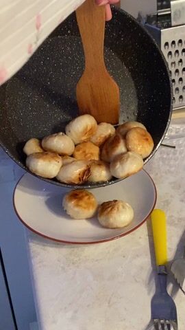 Here is a concise description of the video:

The video shows someone transferring fried dumplings from a non-stick pan to a white plate with a red rim. The person is wearing a white shirt with floral patterns. As the dumplings are being transferred, one falls off the plate and onto the countertop. The background features a kitchen sink, a grater, and a small white bowl. A yellow-handled fork is visible next to the plate of dumplings.
