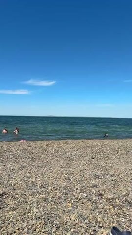 A wide, eye-level shot captures a sunny beach scene in Taman, Russia. The sky is a brilliant, clear blue, with only a few faint, wispy white clouds high near the horizon on the left side, indicating a beautiful day.

The main body of water, likely the Black Sea, displays a greenish-blue hue and appears relatively calm, with small ripples on its surface. Three individuals are visible enjoying the water. Two are closer to the shore, appearing waist or chest deep, with their heads and shoulders above the surface. A third person is further out, almost entirely submerged with only their head visible. A small, pink object, possibly a child's toy or float, is seen floating near the two closer individuals.

The foreground is dominated by a wide expanse of beach composed entirely of smooth, rounded pebbles and small stones in various shades of grey, brown, and off-white. There is no sand visible. The water gently meets the pebbled shore. The bright light suggests it is midday or early afternoon, with perfect weather for a swim and enjoying the coastline.