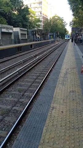 From a standing position on a railway platform, a train approaches along two sets of tracks. The platform has textured gray and yellow tiles, and a dark gray border.  Across the tracks is another station platform with closed buildings. The approaching train is pale blue and white, with some details in black.  Some pedestrians are visible standing on the platform. A few trees and buildings rise in the background. The lighting is bright.