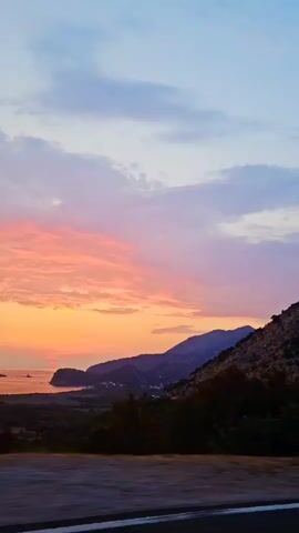 This is a vertical, handheld video of a sunset over a body of water and mountains, likely filmed from a moving vehicle. The sky is filled with clouds in shades of pink, orange, and blue, with the sun a visible orange orb near the horizon. A coastline with mountains and a small village or town is visible in the mid-ground. A road or highway barrier is in the foreground.