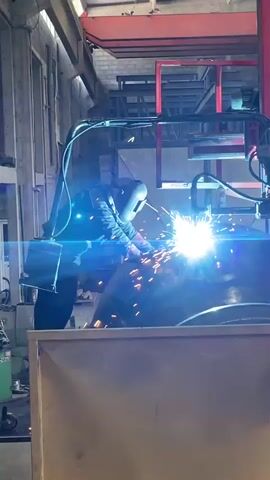 In an indoor setting, a welder wearing protective gear including a face shield and gloves, is engaged in welding a cylindrical object. The welding process produces bright sparks and a vivid light. The welder appears to be standing and working on the object positioned atop a metallic platform. The workshop environment includes equipment like welding machines, tools, and red industrial machinery, all under a high ceiling. The space is well-lit, likely with industrial lighting.