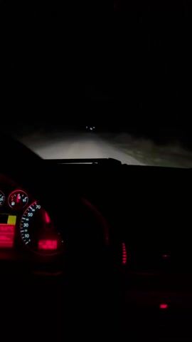 The video shows a dark, in-car view of driving at night. The dashboard and speedometer are visible, illuminated in red, showing the car traveling at approximately 65 km/h. Outside, the road ahead is lit by headlights, with sparse, dark trees lining the sides. No people are discernible, and the focus is on the car's controls and the road ahead.