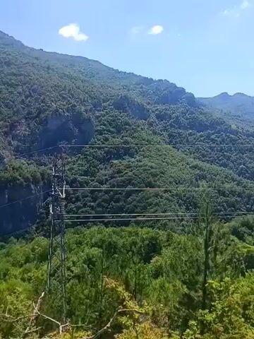 The video shows a panoramic view of a lush, green mountainous landscape under a bright blue sky with scattered white clouds. The camera pans from left to right, revealing rolling hills covered in dense forestation. In the foreground, there are trees and foliage, with some taller pine trees visible. On the left side of the frame, a tall metal electricity pylon with wires stretches across the scene, indicating the presence of human infrastructure within the natural environment. The overall impression is one of natural beauty and expansive wilderness, with a subtle reminder of human presence. There are no people or animals visible in the video.