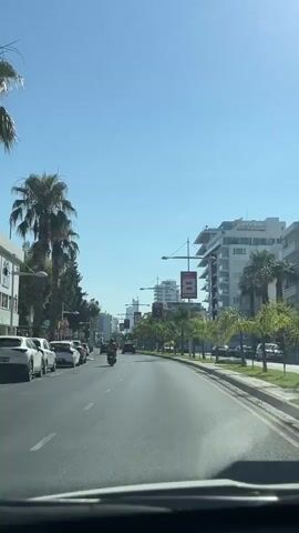 The video shows a POV shot from inside a vehicle moving down a paved road in a city. The sky is clear and blue, suggesting a sunny day. On the left side of the road, there are palm trees, other trees, and parked cars. A motorcycle is visible in the left lane. On the right side of the road, there are more palm trees, sidewalks, and modern buildings. Streetlights with banners attached are spaced along the right side of the road. Several cars and motorcycles are on the road ahead. The vehicle is traveling at a moderate speed, and traffic appears to be light.