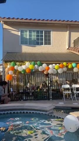 The video shows a backyard party by a pool. A house with a stucco exterior and a tiled roof is visible in the background. A string of colorful balloons is hung across a fence in front of the house. Several inflatable pool floats are in the swimming pool. A person is seen walking towards the pool. Further in the background, a person is standing at a table near a DJ setup. The sky is clear and blue.