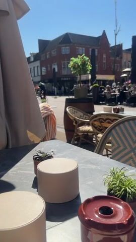 The video shows a sunny outdoor scene from a table at a cafe. A gray, marble-like tabletop is in the foreground, with several small items: two cylindrical objects, a small succulent, a small green plant, and a dark red ashtray. Behind the table are chairs with woven patterns. In the background, across a pedestrian area, is a two-story red brick building with white-framed windows. There are shops visible, including one labeled "PURDEY" and another with "COCKTAILS" on its awning. People are walking and seated at outdoor tables on the other side of the pedestrian area. A person rides a bicycle through the scene. An orange tree in a pot is placed atop a wooden barrel. An umbrella is partially visible to the left of the scene. The sky is clear and blue.