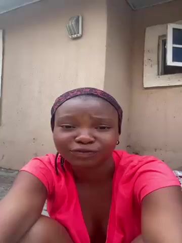 A medium shot shows a woman sitting outside a building. She is wearing a red top and a headband. Her hair is in braids. She is looking directly at the camera and speaking. She sticks out her tongue and then smiles. The building is light brown with two windows. There is a light fixture above her head. The video appears to be filmed outdoors during the day.