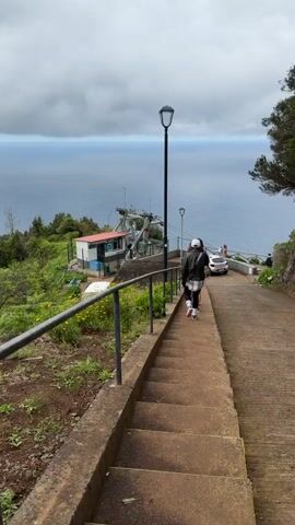 A woman with a white hat, black jacket, and black and white checkered pants walks down a steep, concrete stairway next to a concrete road on a hillside. The road descends toward a viewing area with a white car and several people near a metal railing overlooking the ocean. A small building is to the left of the stairs with a structure for a cable car behind it.  A black metal railing flanks the stairs, and bright green vegetation borders the scene on both sides. The ocean is visible in the background under an overcast sky.