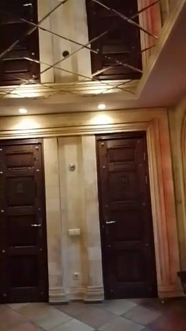 The camera pans up and around a room with a decorative ceiling. The ceiling features a geometric pattern with dark brown panels. A large, ornate chandelier hangs from the center. The walls are finished in a faux stone texture, with two dark brown doors flanking the center. Arched alcoves with decorative shields are visible, as well as red velvet curtains. A dark brown tufted chair is also present. In the background, tables and chairs suggest a dining area.