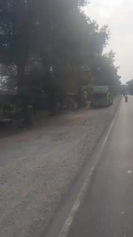 A truck is parked on the side of a dusty road, next to a small structure and under the shade of trees. The truck is green and has a large cargo bed with metal bars. The road is mostly dirt and gravel, with a paved section on the right side. In the distance, more trees and some buildings can be seen. The sky is bright, suggesting it is daytime. The camera appears to be mounted inside another vehicle, possibly a truck or car, as evidenced by the reflection in the side mirror.