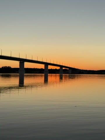 A video depicts a bridge spanning a body of water during sunset. The sky transitions from a light orange near the horizon to a pale gray at the top. The bridge, silhouetted against the colorful sky, has regularly spaced lampposts along its length. Its reflection is visible on the still surface of the water. The water itself is a muted gray, reflecting the colors of the sky and the bridge. A faint silhouette of land and trees is visible in the background on the far side of the bridge. The scene is calm and still, with no visible movement other than the subtle shifts in light as the sun sets.