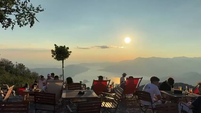 The video captures a scenic view from a mountaintop eatery during sunset. Several people are seated at tables and in lounge chairs, enjoying drinks and conversation. The sun is low in the sky, casting a warm glow over a vast expanse of mountains and a body of water. The atmosphere appears relaxed and social.