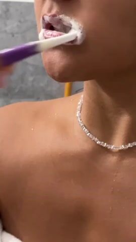 A close-up shot shows a person brushing their teeth. A purple and white toothbrush with white toothpaste is being used to brush the person's teeth. The person's lips are visible, partially covered in toothpaste foam. The person's bare shoulder and neck are also visible, with a delicate, clear beaded necklace around their neck. The background is blurred, with what appears to be a gray tiled wall and a yellow object.