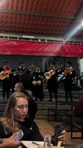 Here's a concise description of the video:

The video shows a group of five musicians performing on a small staircase in an indoor venue, possibly a restaurant. They are all dressed in matching black outfits with decorative red and white elements. Four of them are playing guitars, while the fifth is playing small hand drums. In the foreground, a young woman with long blonde hair is sitting at a table, looking at her phone. Other diners are visible in the background. The venue has a red-tiled roof and is lit with artificial lights.