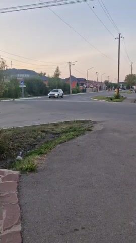 The video shows a street scene with a car driving on the road. There are power lines overhead and houses in the background. The sky is overcast. The time of day appears to be either early morning or late afternoon, given the lighting. There is a pedestrian crossing sign on the left side of the road. A white car is visible in the distance, approaching the intersection. The road surface is asphalt. There is some grass and dirt on the side of the road.
