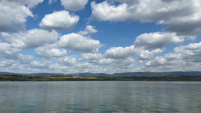 The video shows a wide, calm body of water under a bright blue sky dotted with fluffy white clouds. The horizon line is marked by a low, undulating landscape of hills covered in sparse vegetation, with a few distant structures that appear to be industrial or infrastructure-related on the left side. The water's surface is slightly rippled, reflecting the sky and clouds in a muted, greenish-blue tone. There are no people or obvious animals visible in the frame. The scene suggests a natural outdoor environment, possibly a reservoir or a large lake, on a partly cloudy day. The camera appears to be positioned low, near the water's surface, and is slowly panning across the scene from left to right.