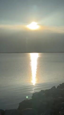 A vertical, eye-level shot of a body of water during sunset. The sun is partially obscured by clouds, casting a bright, golden reflection on the water's surface. The water is calm with gentle ripples. In the foreground, rocks and some dry vegetation are visible along the shoreline. The sky is hazy with rays of sunlight piercing through.