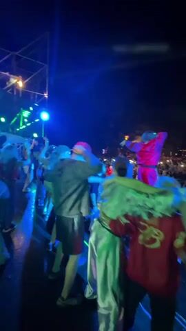 The video shows a nighttime outdoor concert. A performer in a bright red outfit is on stage, facing a large crowd. Several people, appearing to be friends, are gathered together on what looks like a raised platform or the edge of the stage, with their arms around each other. They are all facing the performer and the crowd. The lighting is predominantly blue and some other colored stage lights. The crowd is visible in the background, with many holding up their phones. The overall atmosphere suggests a lively and energetic event.