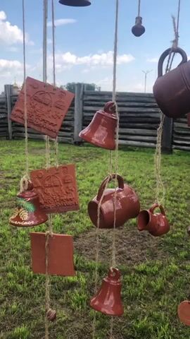 The video shows a collection of clay pottery items hanging from strings against a blue sky with clouds. The items include bells, mugs, and flat decorative pieces with relief designs. The pottery is reddish-brown in color and appears to be handmade. In the background, there is a wooden fence and green grass. The overall setting suggests an outdoor market or craft fair. There are no people visible in the video, and no actions or interactions are taking place beyond the gentle movement of the hanging pottery in what seems to be a light breeze.