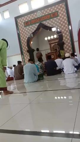 The video shows the interior of a mosque where men are seated on the floor facing the front. A man stands in front of them behind a dark-colored archway with a decorative panel above it. The men are wearing a variety of clothes, some white and some of other colors. The floor is tiled with light-colored tiles. There are windows near the top of the wall.