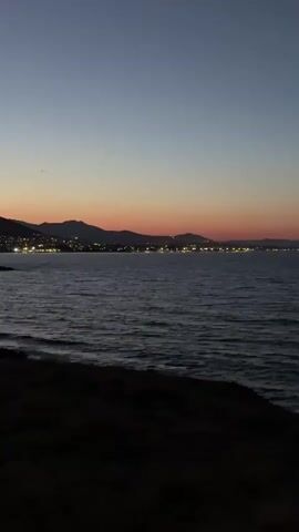 The video shows a wide shot of a calm ocean during twilight. The sky transitions from a deep blue at the top to hues of orange and pink near the horizon, indicating either sunrise or sunset. In the distance, a coastline is visible with mountains behind it. A line of lights, likely from a town or city, stretches along the coast. The water is dark and shows gentle ripples. The foreground is dark, suggesting it is either a beach or a cliff overlooking the water, with some indistinct dark shapes that could be rocks or vegetation. There are no people or specific objects clearly identifiable in the foreground or the water. The overall atmosphere is serene and peaceful.