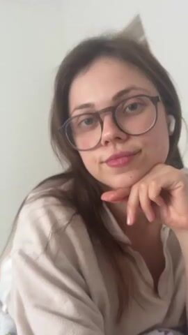 A woman with long brown hair and glasses, wearing earbuds and a beige top, is looking downward with a smile and her hand under her chin. She looks like she is watching something. The camera turns to show a tablet laying on a bed with a white floral patterned bedspread. The tablet screen displays a man in green medical scrubs speaking. The captions at the bottom of the screen state "Trauma, oncology, cardiology and pediatrics all need consults."