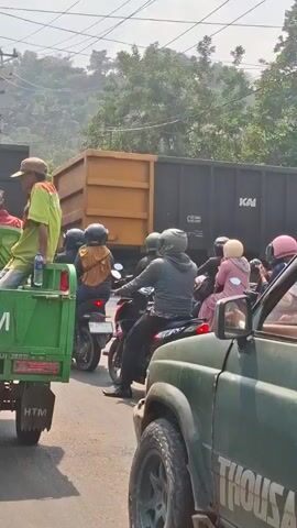 A freight train is passing through a street crossing, causing a traffic jam. Several people on motorcycles are waiting for the train to pass. The train cars are dark gray with white lettering indicating "KAI" and other numbers. Some of the motorcycles have passengers. A green truck is visible in the foreground on the left, and a dark green SUV is in the foreground on the right. Trees and power lines are visible in the background. The sky is clear and bright.