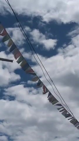 The video shows a string of prayer flags strung between two points against a backdrop of a blue sky with white clouds. The flags are small and rectangular, in various colors including blue, white, red, green, and yellow, and are strung together on a wire or rope. The flags are blowing gently in the wind. The perspective is looking upwards towards the sky.