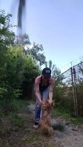 A woman wearing ripped jeans, a black crop top, a black baseball cap, and black slides is on a dirt path outdoors, flanked by green foliage on the left and a barbed wire fence on the right, under a clear blue sky. She bends down to pick up a small, light brown dog. She then lifts the dog to face level, kisses its head, and lowers it.