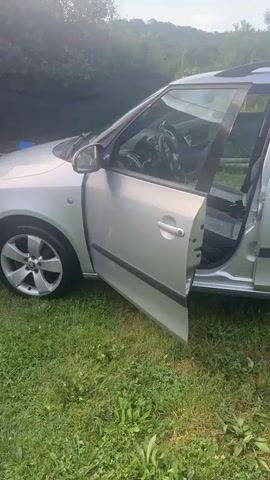 The video shows a silver car parked on a grassy area next to a campsite. The car's front passenger door is open. Behind the car is a tent and a white shed-like structure. The camera then pans to the right, revealing a dog standing on a concrete patio under a shaded structure with a table and chairs. The lighting is bright and sunny.