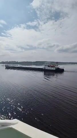 The video shows a large barge with a white and black tugboat on the back moving along a wide, calm body of water, likely a river or lake. The barge appears to be carrying a flat load, and the water is dark with subtle ripples reflecting the bright sunlight. The sky is partially cloudy with a mix of white and gray clouds. The camera seems to be positioned on another boat or structure near the water's edge, as part of a white surface is visible in the foreground. In the distance, the shoreline features some greenery and buildings.